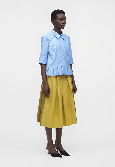 Femme portant une chemise à rayures bleu clair avec boutons et plis, et une jupe midi plissée jaune, debout avec des talons pointus noirs.