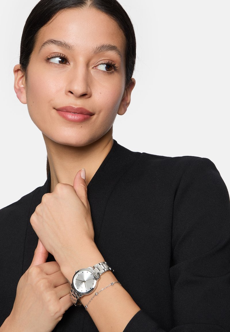 Zilveren metalen horloge met een ronde wijzerplaat, schakelsband en minimalistisch ontwerp. Draagt op een pols met een delicaat zilveren armband. Zwarte kleding achtergrond.