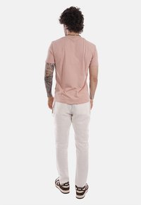 Homme avec des tatouages sur le bras gauche, portant un t-shirt à manches courtes rose clair, un pantalon blanc et des baskets Nike noires et blanches, vu de dos sur un fond uni.