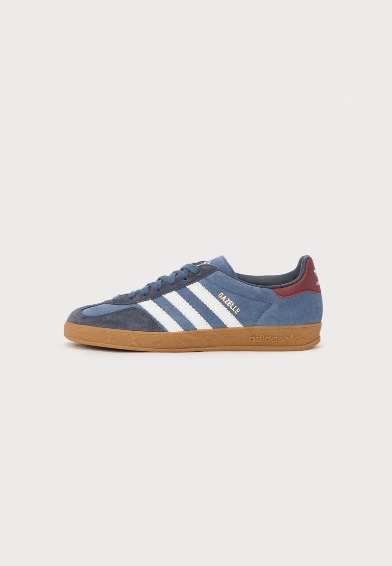 Baskets Adidas Gazelle bleues avec des bandes blanches, une semelle en gomme et un renfort de talon bordeaux, vue latérale sur fond blanc.