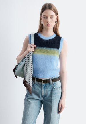 Jeune femme portant un haut sans manches bleu à rayures pelucheux et un jean bleu clair, tenant un sac bleu clair avec une écharpe à motifs.