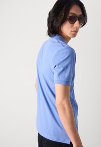 Camiseta polo de culoare albastru deschis, dintr-un material de bumbac moale, cu guler clasic și mâneci scurte, prezentată din spate, cu un design simplu.