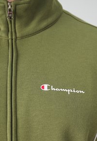Zelená zipová bunda vyrobená z mäkkého materiálu, s vysokým golierom, čiernym zipsom a bielym logom "Champion" s červeným akcentom.