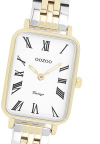 Montre rectangulaire en métal argenté et doré avec un cadran blanc, des chiffres romains noirs et des aiguilles dorées. Dispose d'une couronne sur le côté droit.