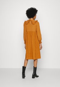 Vestido ankle-length em tom rust, com tecido texturizado, gola com volantes e mangas longas, combinado com botas de salto pretas.