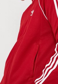 Punainen urheilutakki, jossa on vetoketjukiinnitys, valkoiset korostusraidat hihoissa ja pieni logo rinnassa. Sileä rakenne.