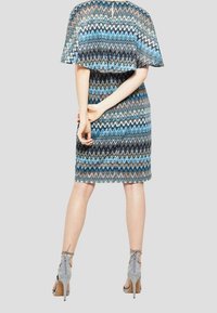 Robe mi-longue avec un motif zigzag en bleu, noir et blanc. Dotée d'un haut en forme de cape et d'une fermeture éclair au dos. Associée à des sandales à talons hauts grises.