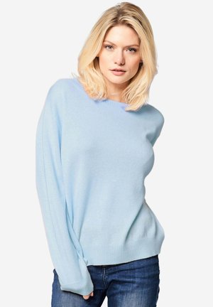 Femme blonde en pull bleu clair à manches longues et jean bleu foncé posant devant un fond blanc, regardant vers la caméra.