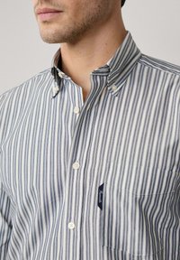 Camisa de botones con rayas verticales en azul marino y blanco, fabricada en algodón, con cuello puntiagudo, bolsillo en el pecho y una etiqueta de marca.