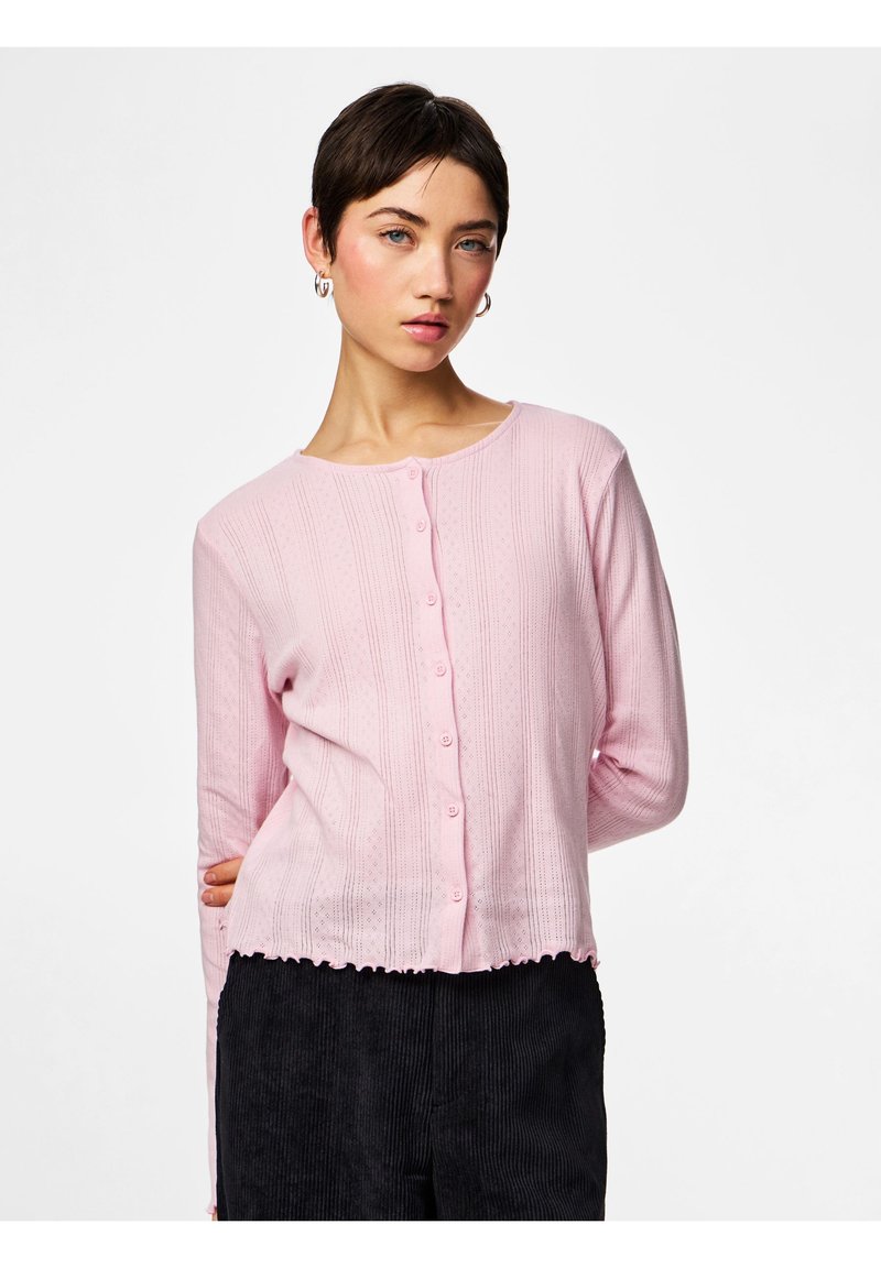 Νέα γυναίκα με κοντά σκούρα μαλλιά, ντυμένη με ροζ ανοιχτό πλεκτό ζακετάκι με κουμπιά και μαύρο παντελόνι, ποζάροντας μπροστά σε λευκό φόντο.