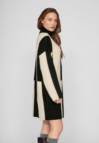 Adīta džempera jaka ar melnbaltiem vertikāliem svītrām, garām piedurknēm un augstu apkakli, ar teksturētu mīkstu apdari un ceļu garumu.