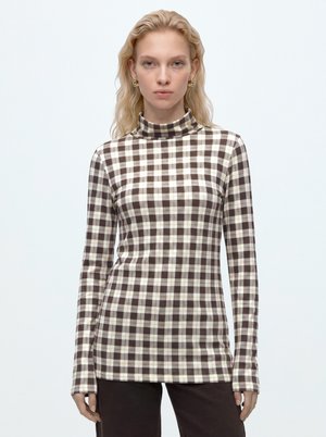 Blonde woman wearing a brown and white checkered long-sleeve turtleneck top and dark brown pants, standing against a light background.