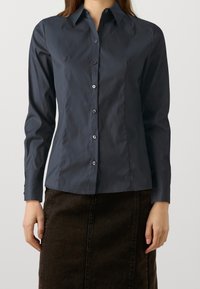 Donna che indossa una camicia a maniche lunghe di bottoni blu scuro aderente e una gonna marrone scuro su uno sfondo neutro.