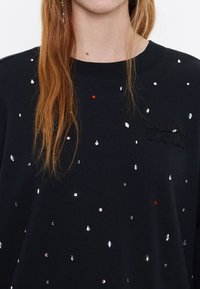 Sudadera negra con diversas formas de lentejuelas en plata, rojo y rosa. Tiene un cuello redondo y un logo texturizado en la parte frontal.