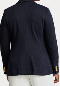 Blazer azul marino con un tejido texturizado, abertura trasera sencilla, dos bolsillos laterales y detalles de botones dorados en los puños.