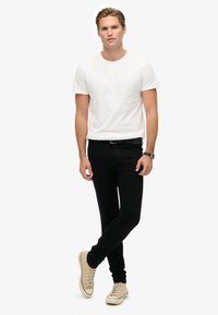 Camiseta blanca de cuello redondo, jeans ajustados negros, zapatillas beige con diseño de cordones, en pose de pie frente a un fondo blanco.