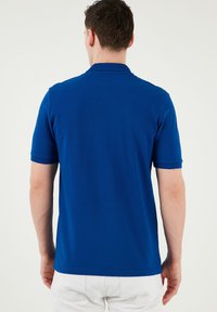 Zila kokvilnas polo krekls ar īsām piedurknēm, teksturētu apkakli un gludu virsmu. Nav redzamu logo vai rakstu.