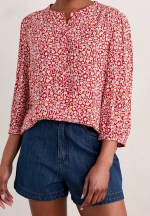 Person trägt eine rote, geblümte, langärmelige Bluse mit Knöpfen und dunkelblaue Jeansshorts.