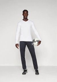 Λευκό αθλητικό μπλουζάκι με μακριά μανίκια και μαύροι αθλητικοί παντελόνες, κρατώντας μια ποδοσφαιρική μπάλα με σχέδιο. Μαύρα παπούτσια συμπληρώνουν την αθλητική εμφάνιση.