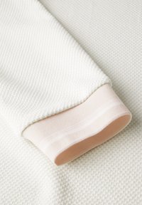 Weißer strukturiertes Gewebe mit einem pfirsichfarbenen gerippten Bündchen. Das Material weist ein gewebtes Muster auf, das Tiefe und visuelles Interesse verleiht.