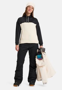 Zwarte en crèmekleurige pullover met lange mouwen en een gestructureerd ontwerp. Zwarte skibroek en een witte jas. Houdt skibril vast met een kleurrende lens.