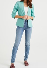 Blazer verde agua con mangas de tres cuartos, combinado con jeans ajustados azul claro y zapatos tipo slip-on burdeos. Top de encaje blanco visible por debajo.