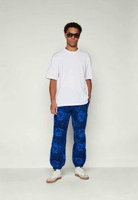 Homem a usar óculos de sol pretos, camisa branca simples de manga curta, calças azuis com padrão floral e ténis brancos, de pé contra um fundo claro.