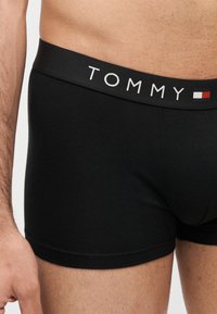 Κοντινό πλάνο στο κάτω μέρος της κοιλιάς ενός άνδρα που φοράει μαύρα μποξεράκια με τη λέξη «TOMMY» και ένα κόκκινο, άσπρο και μπλε λογότυπο στη ζώνη μέσης.