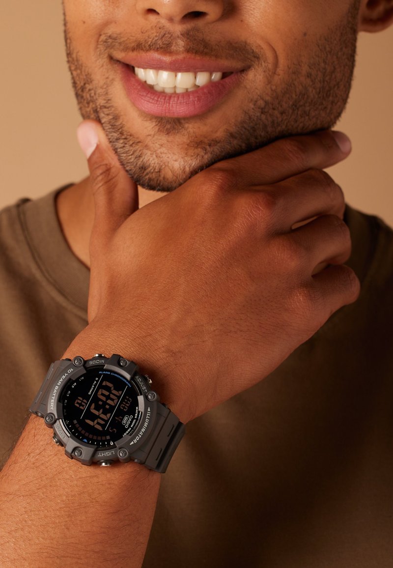 Montre numérique avec un bracelet en silicone gris, cadran noir rond et affichage numérique blanc contrastant. Présente des accents métalliques et des boutons.