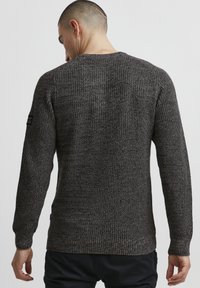 Dark gray knit sweater with a textured finish, crew neckline, and fitted long sleeves. Features a small black branded patch on the sleeve.