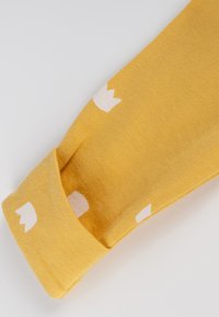 Manica gialla in cotone con motivi a corona color crema chiaro. Presenta un polsino a costine e una texture liscia, evidenziando la semplicità nel design.