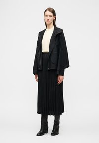 Femme portant une veste noire à capuche, un chemisier crème, une jupe midi plissée noire et des bottes à talons noires, debout devant un fond blanc uni.