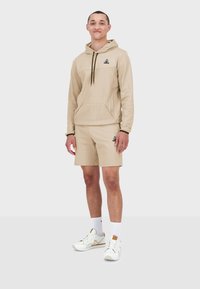 Sudadera beige con bolsillo canguro y logo negro, combinada con pantalones cortos a juego. Lleva zapatillas blancas y calcetines blancos.