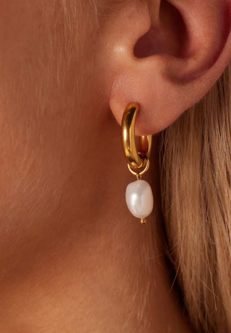 Gouden ringoorbel met een witte, ovale pareldruppel. De gestructureerde gouden afwerking en het hangende ontwerp benadrukken de glans van de parel.