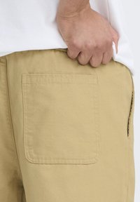 Pantaloncini color cachi con una tasca posteriore semplice e cuciture visibili. Il tessuto appare resistente e strutturato. Mano appoggiata sulla vita.