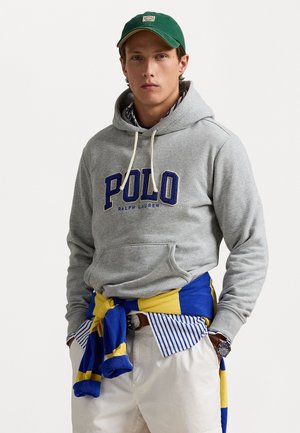 Felpa grigia con logo "POLO", con cordini beige, indossata sopra un maglione a righe blu e giallo legato in vita, abbinata a pantaloni chiari.
