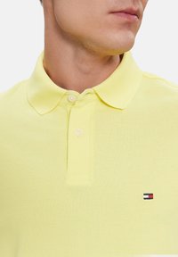 Polo en amarillo claro con un tejido texturizado, dos botones, cuello puntiagudo y un pequeño logo en rojo, blanco y azul en el pecho.
