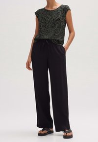 Pantaloni neri larghi con tasche laterali, abbinati a una maglietta nera a maniche corte con un motivo a pois colorati. Sandali neri piatti.