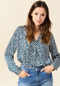 Navyblaue Bluse mit weißem abstraktem Muster, knöpfbare Vorderseite, lange Ärmel und Kragen; kombiniert mit hoch geschnittenen Jeans.