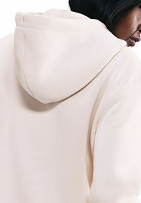 Felpa leggera beige con una consistenza morbida, grande cappuccio e cuciture visibili sul retro, con una vestibilità rilassata.