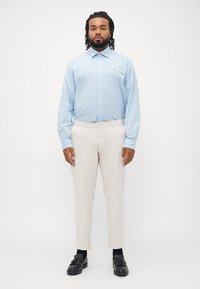Hombre con rastas vistiendo una camisa de botones azul clara, pantalones beige, calcetines negros y mocasines con borlas negros, de pie contra un fondo blanco.