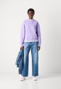 Felpa di cotone viola con polsini a costine, abbinata a jeans in denim blu a gamba larga. Sneaker bianche e una giacca di denim sono tenute in una mano.