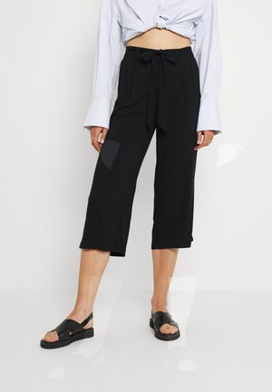 Persoon die zwarte, hoge taille, wijde cropped broek draagt met een gestrikte riem, wit overhemd met lange mouwen gestrikt in de taille en zwarte open-toe sandalen.