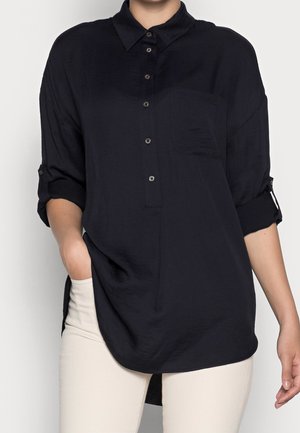 Marineblaue Button-Down-Bluse mit hochrollbaren Ärmeln, einer einzelnen Brusttasche und einem längeren hinteren Saum, aus leichtem, strukturiertem Stoff.