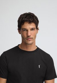 Svart bomulls-t-shirt med rund halsringning och korta ärmar, med en liten vit logotyp på vänster bröst. Mjuk textur.