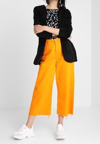 Sort, fluffy jakke, mønstret sort og hvid top, klare orange brede cropped bukser og hvide sneakers med chunky såler.