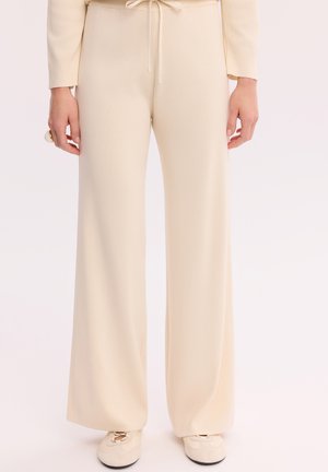 Kobieta w kremowych szerokich spodniach z sznurkiem w talii i dopasowanych kremowych butach, stojąca na prostym jasnym tle.