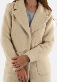 Cappotto in misto lana beige con finitura testurizzata, con rever a punta e due tasche frontali, caratterizzato da una vestibilità comoda e maniche lunghe.