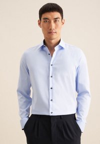Camicia azzurra chiara con colletto strutturato, bottoni di una tonalità più scura e maniche lunghe. Il materiale appare liscio con una leggera lucentezza.