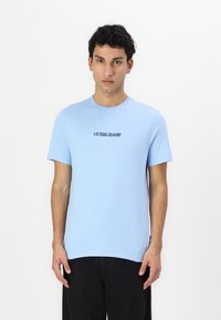 Lühikeste varrukatega, ümarate kaelustega sinine t-särk, mis on valmistatud puuvillast; keskel rinnal on must "GUESS JEANS" logo; sile tekstuur.